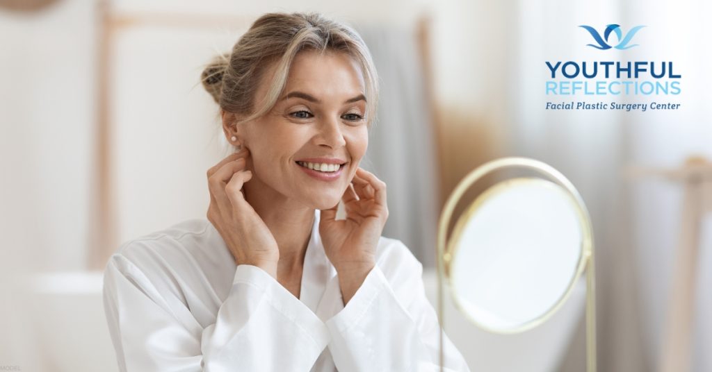 Woman is smiling while looking at reflection in a mirror (model) Youthful Reflections: Michael A. Boggess, MD Logo
