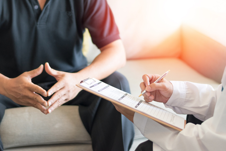 Patient sitting during consultation with doctor taking notes on clipboard.