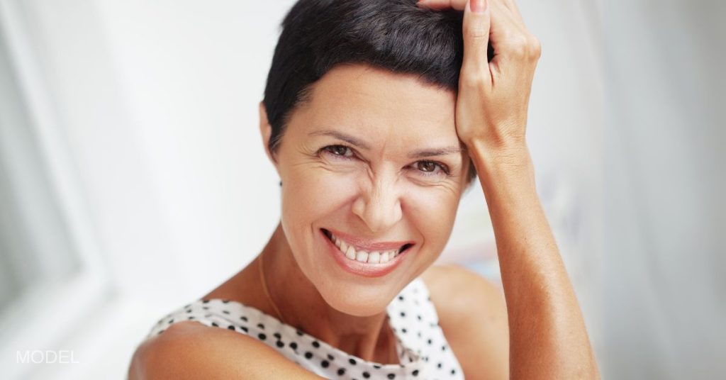 Beautiful middle-aged woman rests her head against her palm while smiling at the camera. (Model)