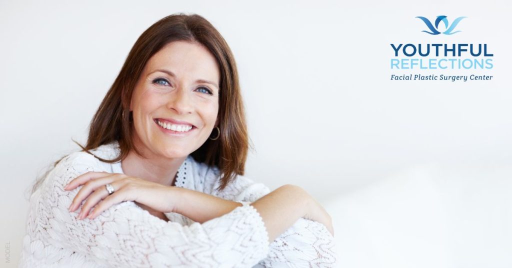 A mature woman smiles gently, with her arms lightly crossed over her chest. (MODEL). Youthful Reflections logo.